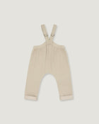 A pair of baby overalls in a light cream or beige color, featuring adjustable shoulder straps with metal rings and rolled cuffs at the bottom, shown flat against a light gray background.