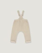 A pair of baby overalls in a light cream or beige color, featuring adjustable shoulder straps with metal rings and rolled cuffs at the bottom, shown flat against a light gray background.