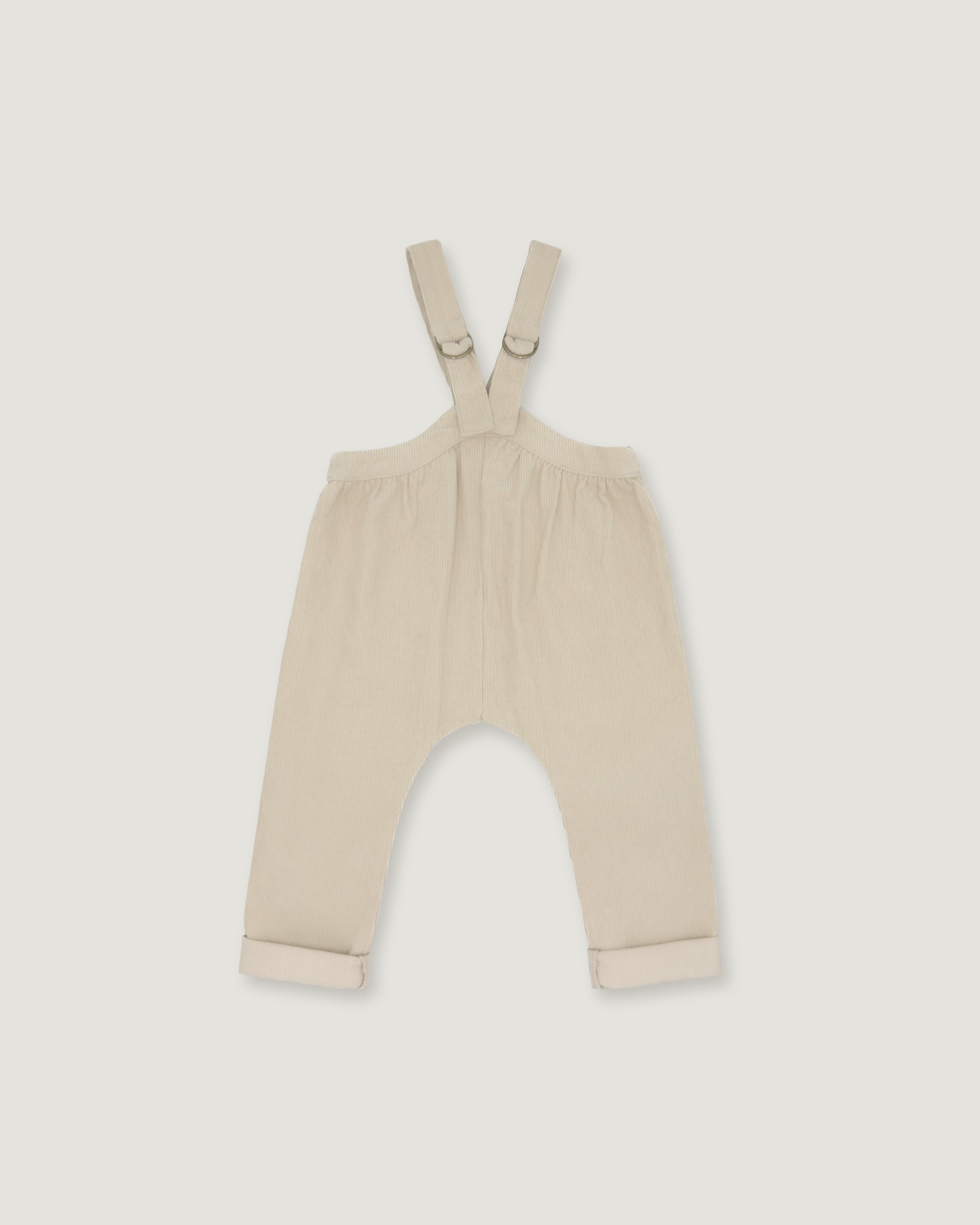 A pair of baby overalls in a light cream or beige color, featuring adjustable shoulder straps with metal rings and rolled cuffs at the bottom, shown flat against a light gray background.