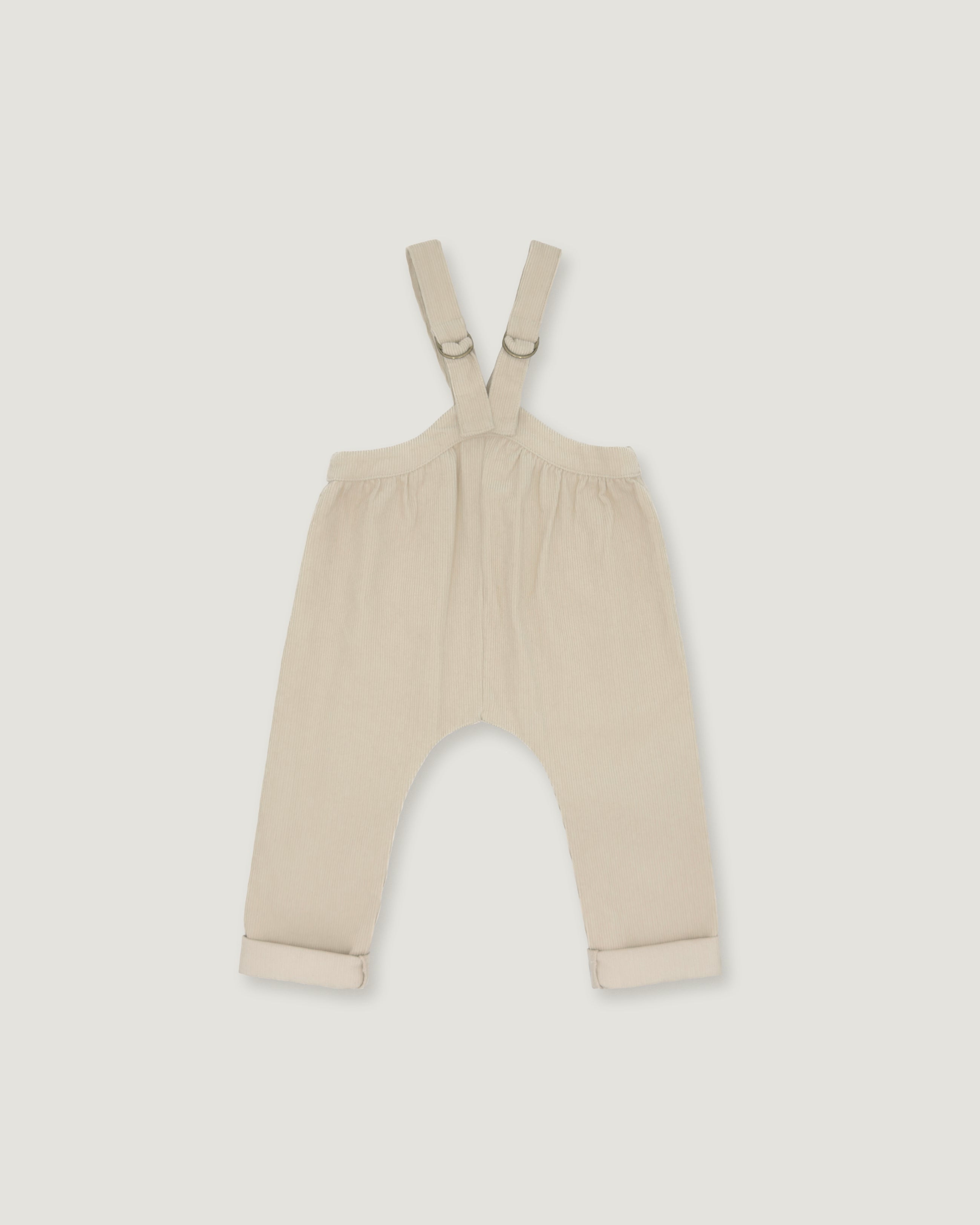 A pair of baby overalls in a light cream or beige color, featuring adjustable shoulder straps with metal rings and rolled cuffs at the bottom, shown flat against a light gray background.