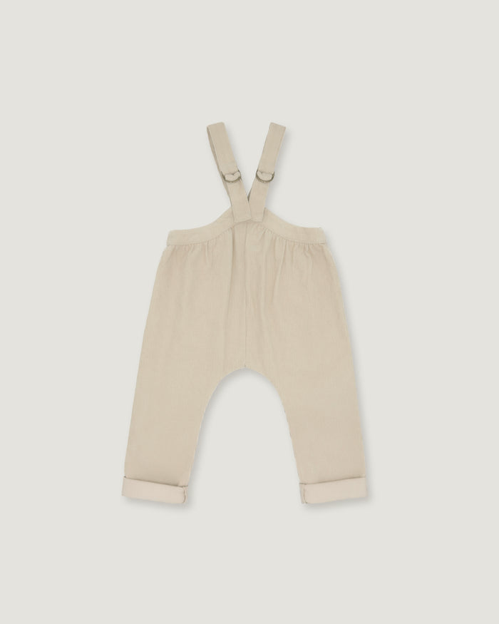 A pair of baby overalls in a light cream or beige color, featuring adjustable shoulder straps with metal rings and rolled cuffs at the bottom, shown flat against a light gray background.