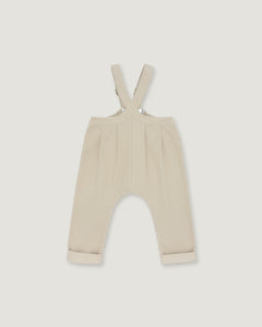 A pair of baby trousers in a light cream or beige color, featuring a bib-style waistband with adjustable shoulder straps and cuffed legs, shown flat against a light gray background.