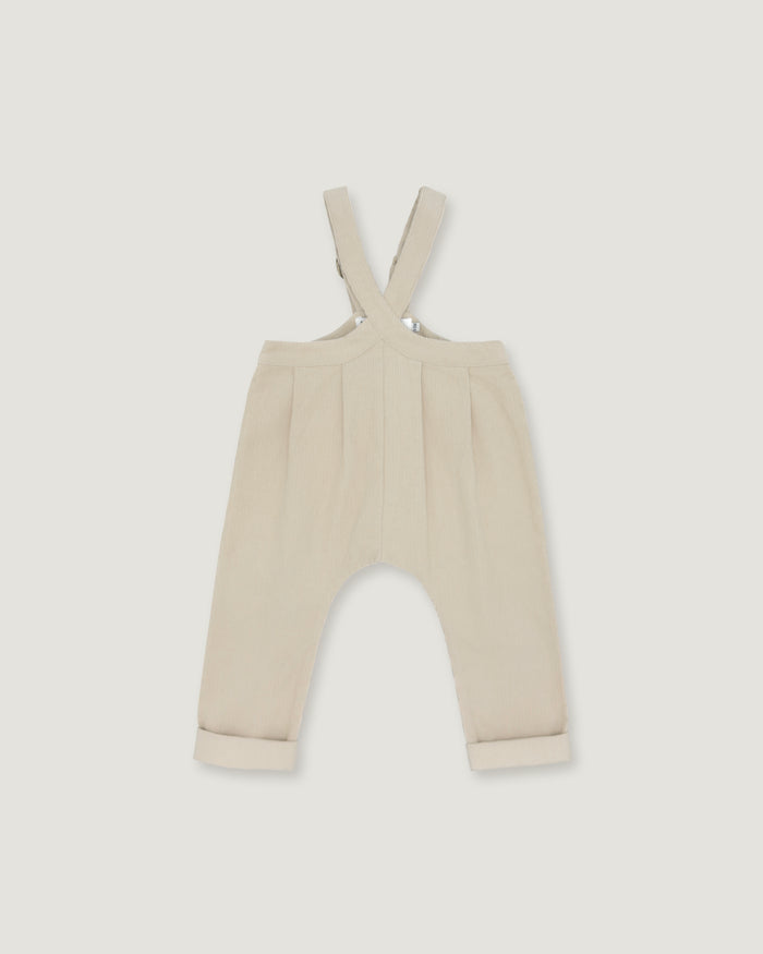 A pair of baby trousers in a light cream or beige color, featuring a bib-style waistband with adjustable shoulder straps and cuffed legs, shown flat against a light gray background.