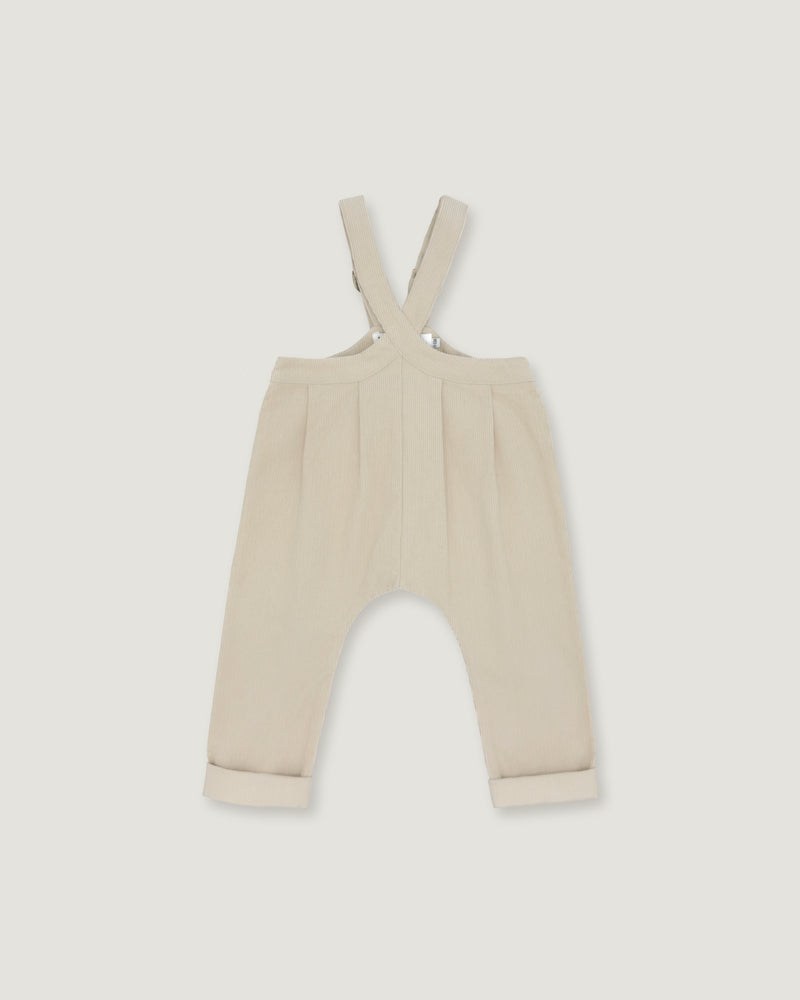 A pair of baby trousers in a light cream or beige color, featuring a bib-style waistband with adjustable shoulder straps and cuffed legs, shown flat against a light gray background.