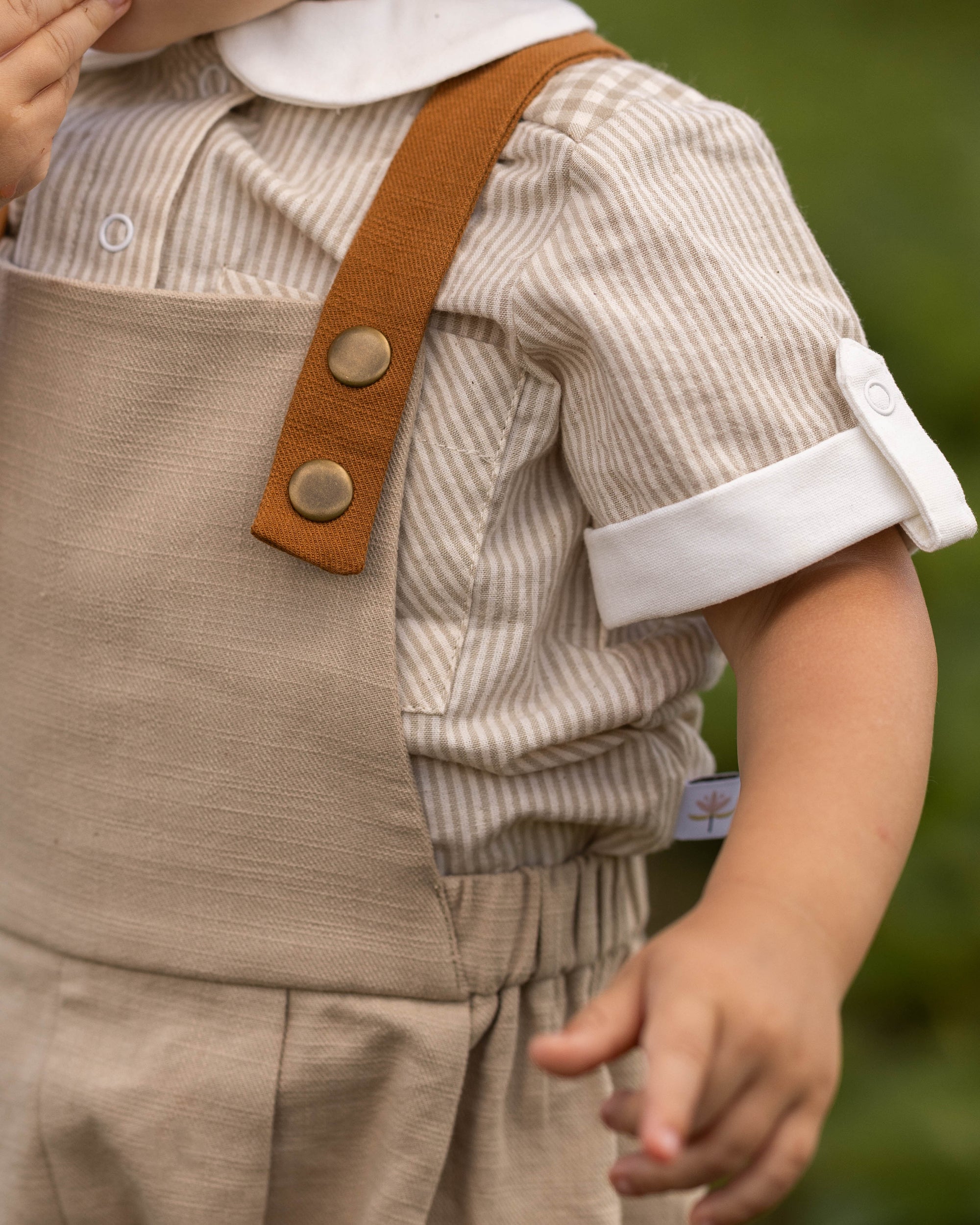 Close-up of a child wearing a beige corduroy overall-style romper with brown shoulder straps and a striped shirt, photographed outdoors against a blurred green background.
