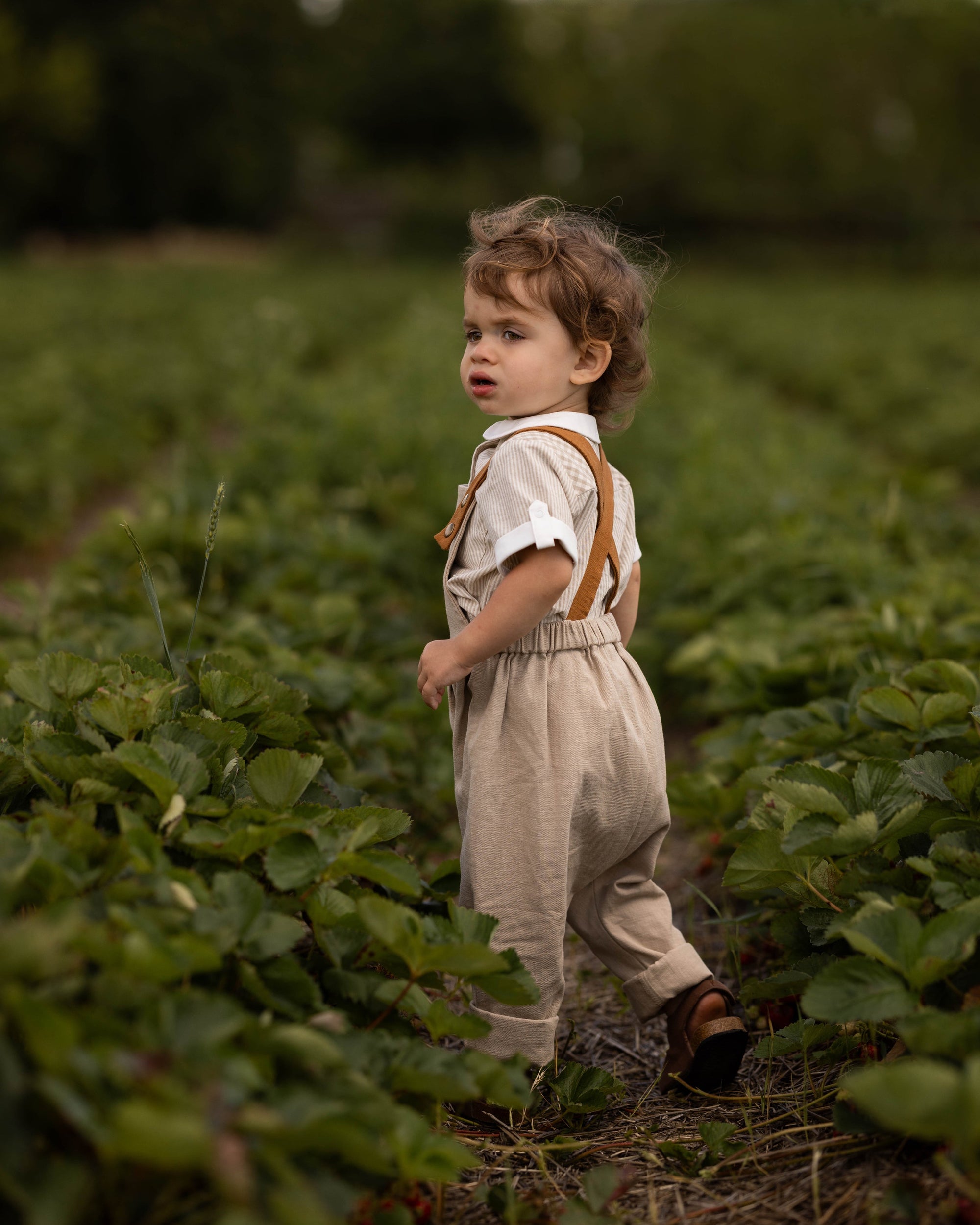 A toddler standing in a green field, wearing a light beige overall-style romper with brown suspenders and a white short-sleeve shirt.