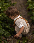 A toddler crouches in a garden path between rows of green strawberry plants, wearing a cream ribbed short-sleeve shirt with brown suspenders and beige trousers.