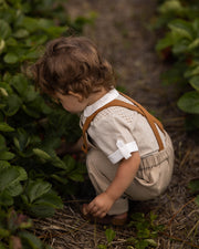 A toddler crouches in a garden path between rows of green strawberry plants, wearing a cream ribbed short-sleeve shirt with brown suspenders and beige trousers.