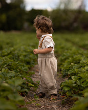 A toddler standing in a field of green plants, wearing a light beige short-sleeve shirt with brown suspenders and matching light beige pants.