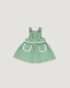 A sage green baby dress with white trim details, featuring a flared skirt and front pockets, shown against a solid light gray background.