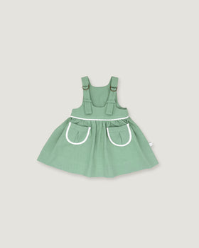A sage green baby dress with white trim details, featuring a flared skirt and front pockets, shown against a solid light gray background.