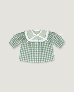 A baby girl's short-sleeve button-up shirt in a green and white gingham check pattern, featuring a wide white ruffled collar with a white cross-front bow, displayed flat against a light beige background.