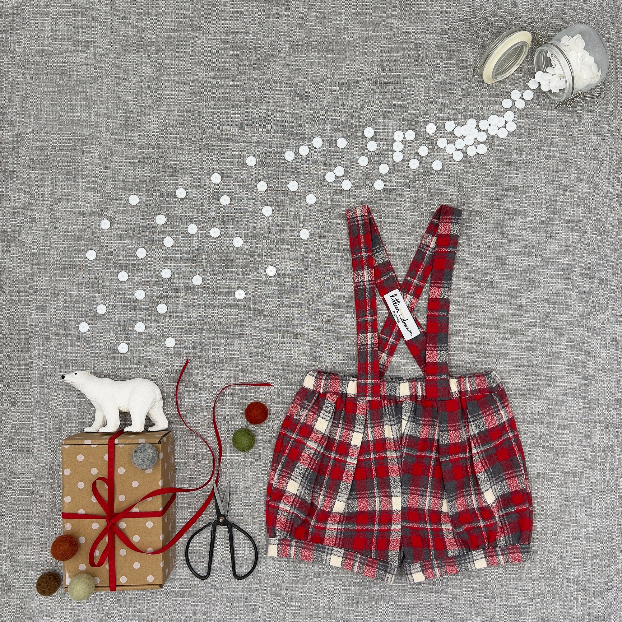 A baby boy's red and beige plaid romper with suspenders, styled in a flat-lay arrangement on a gray linen background with decorative props including a gift box, scissors, a jar of white dots, and a small polar bear figurine.