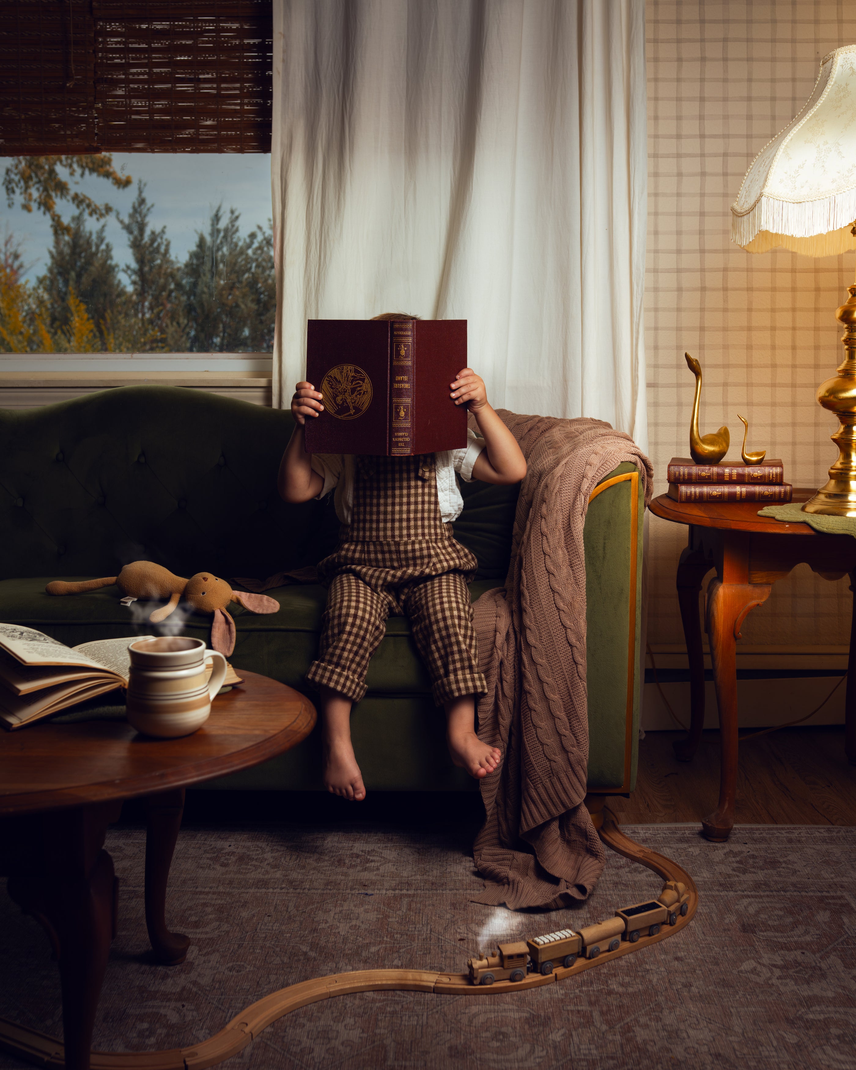 A child in brown plaid pajamas sits on a dark green sofa in a cozy, warmly lit room, holding a dark red book to their face. A toy train tracks a path across the floor in front of the sofa, with a lamp, side table, and window in the background.