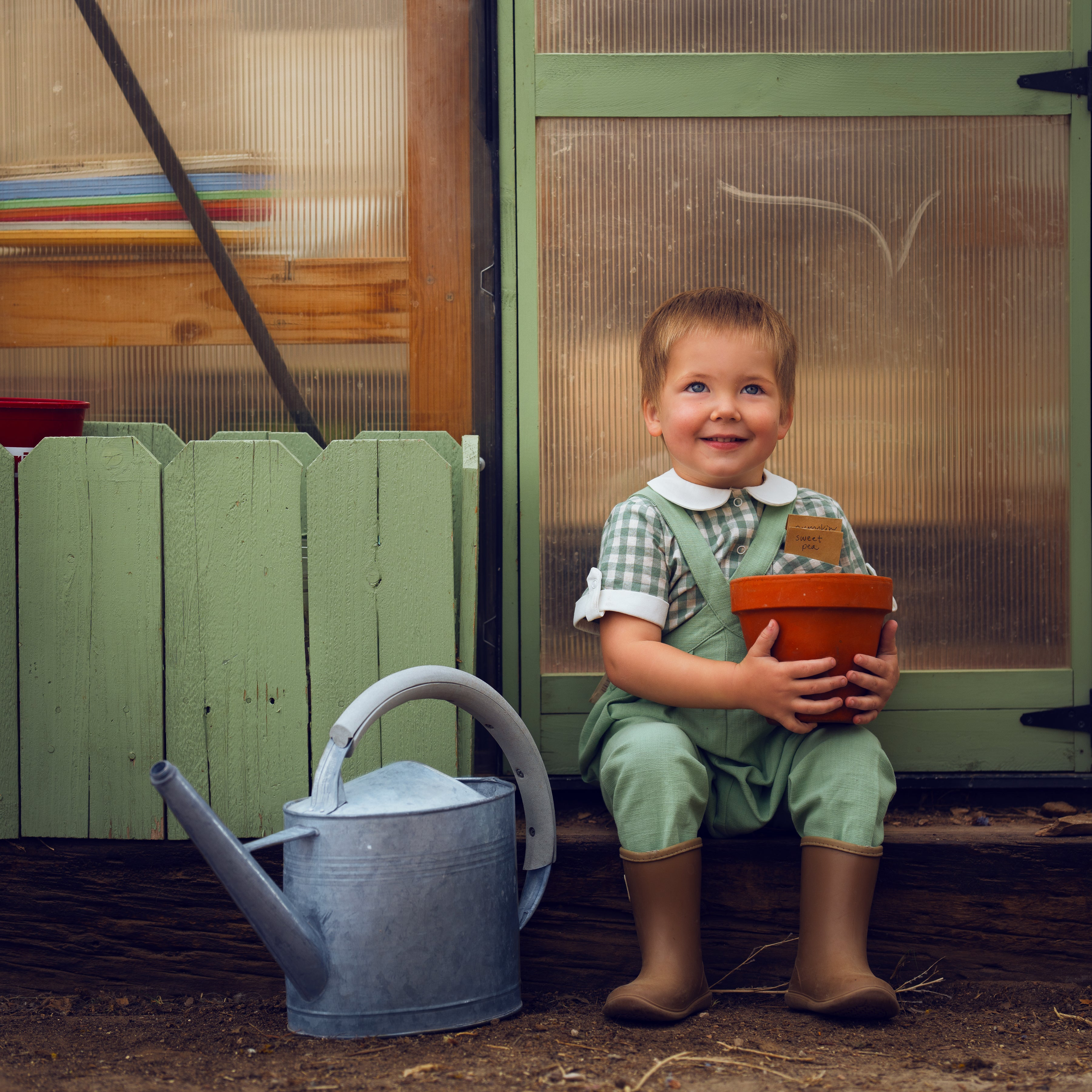 Child holding a pot and smiling in front of a green door with a watering can on the ground.