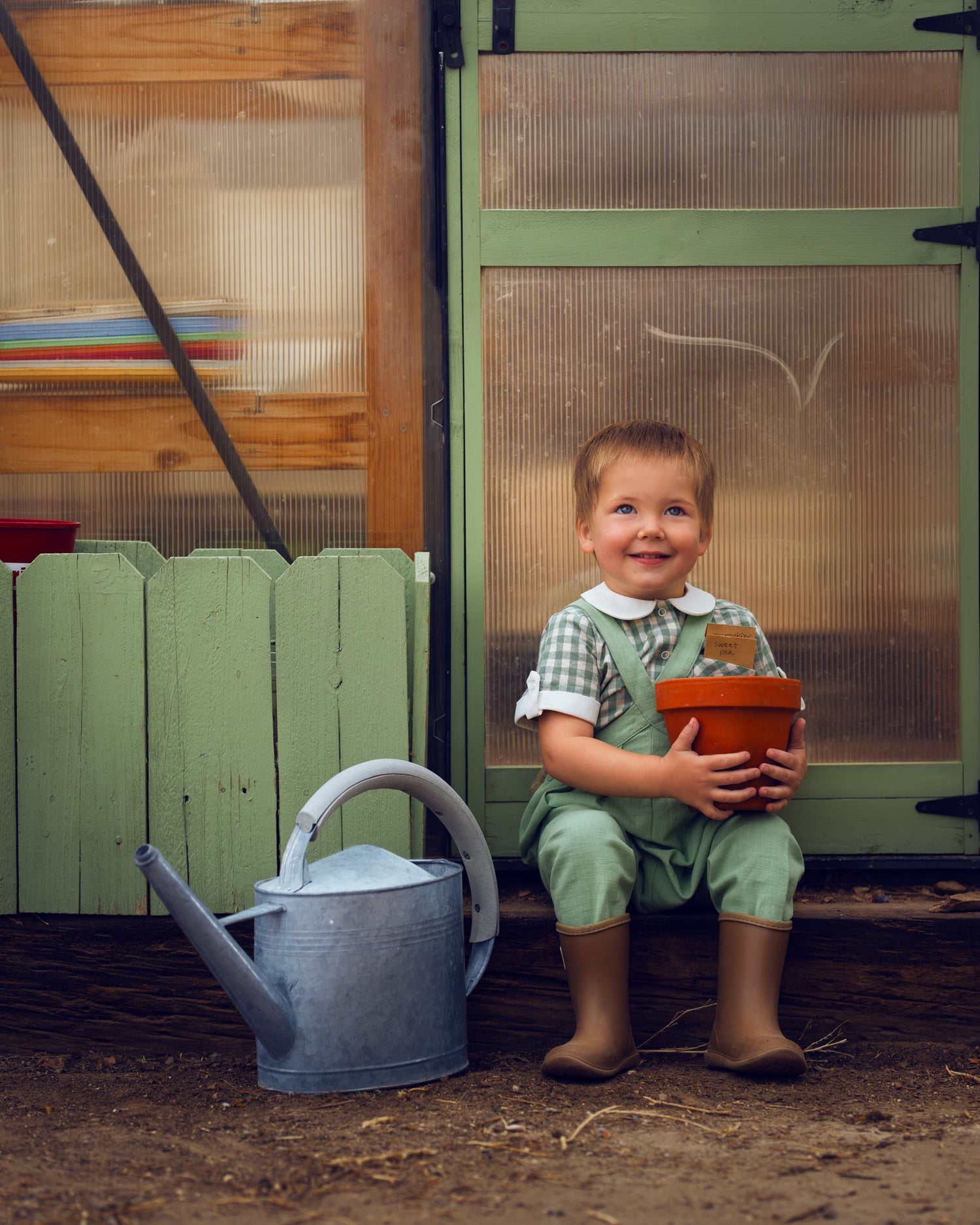 Child holding a pot and smiling in front of a green door with a watering can on the ground.