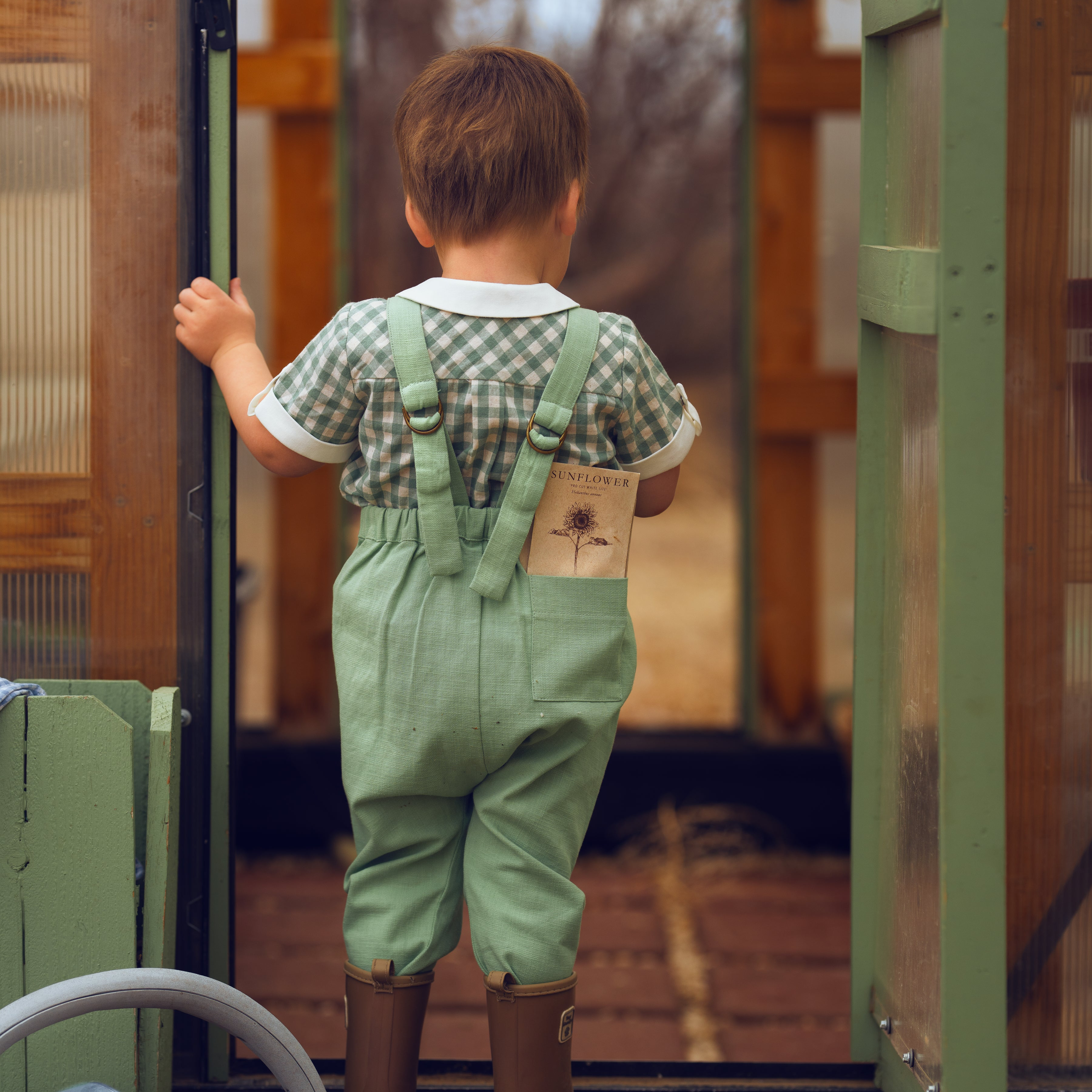 Child wearing green overalls and brown boots standing inside a wooden shed.