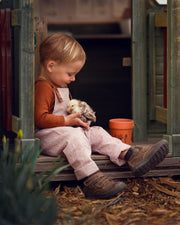 Little boy sitting in checkered brown and white linen overalls playing with chicks in a barn.