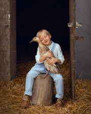 A young boy in blue gingham overalls and a white shirt sits on a wooden stump in a barn, holding a brown duck. The scene is filled with straw on the floor and a dark interior behind them.