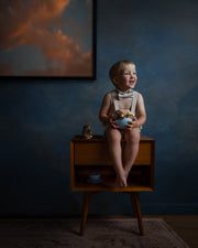 A toddler boy wearing a cream suspender outfit and bow tie sits on a mid-century modern wooden side table, holding a blue bowl. The scene is set against a dark blue textured wall with a framed sunset painting in the background.