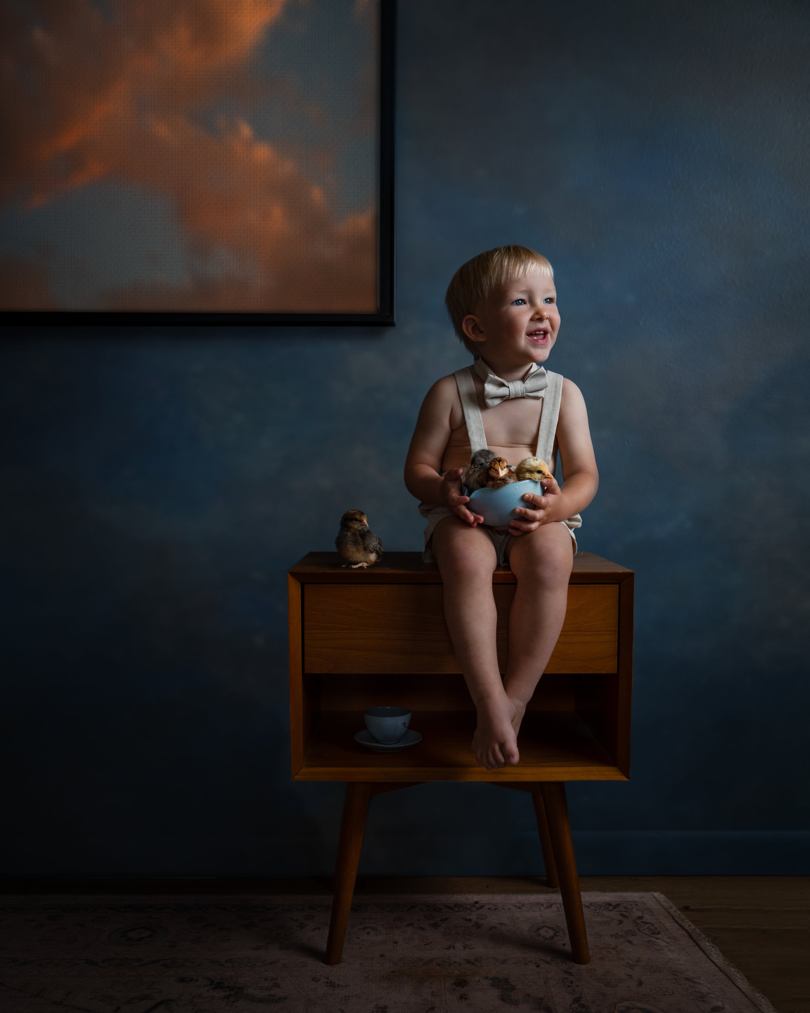 A toddler boy wearing a cream suspender outfit and bow tie sits on a mid-century modern wooden side table, holding a blue bowl. The scene is set against a dark blue textured wall with a framed sunset painting in the background.