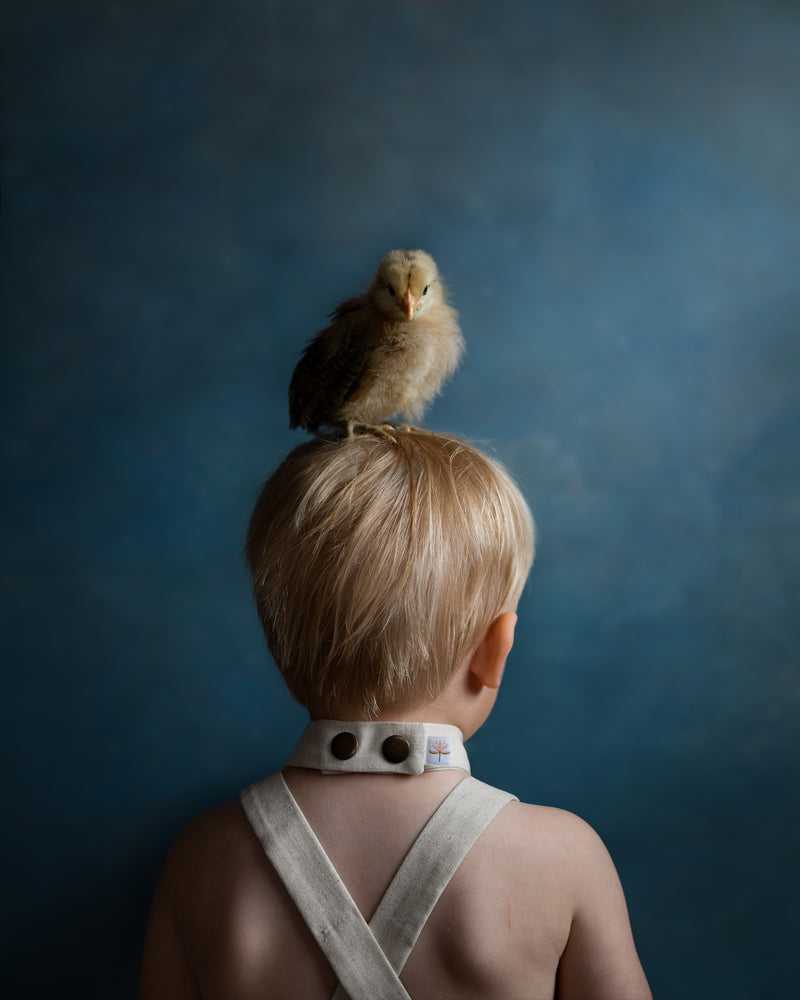 A young child seen from behind wearing a white neck brace, with a small fluffy chick perched on the child's head, set against a dark blue textured studio background.