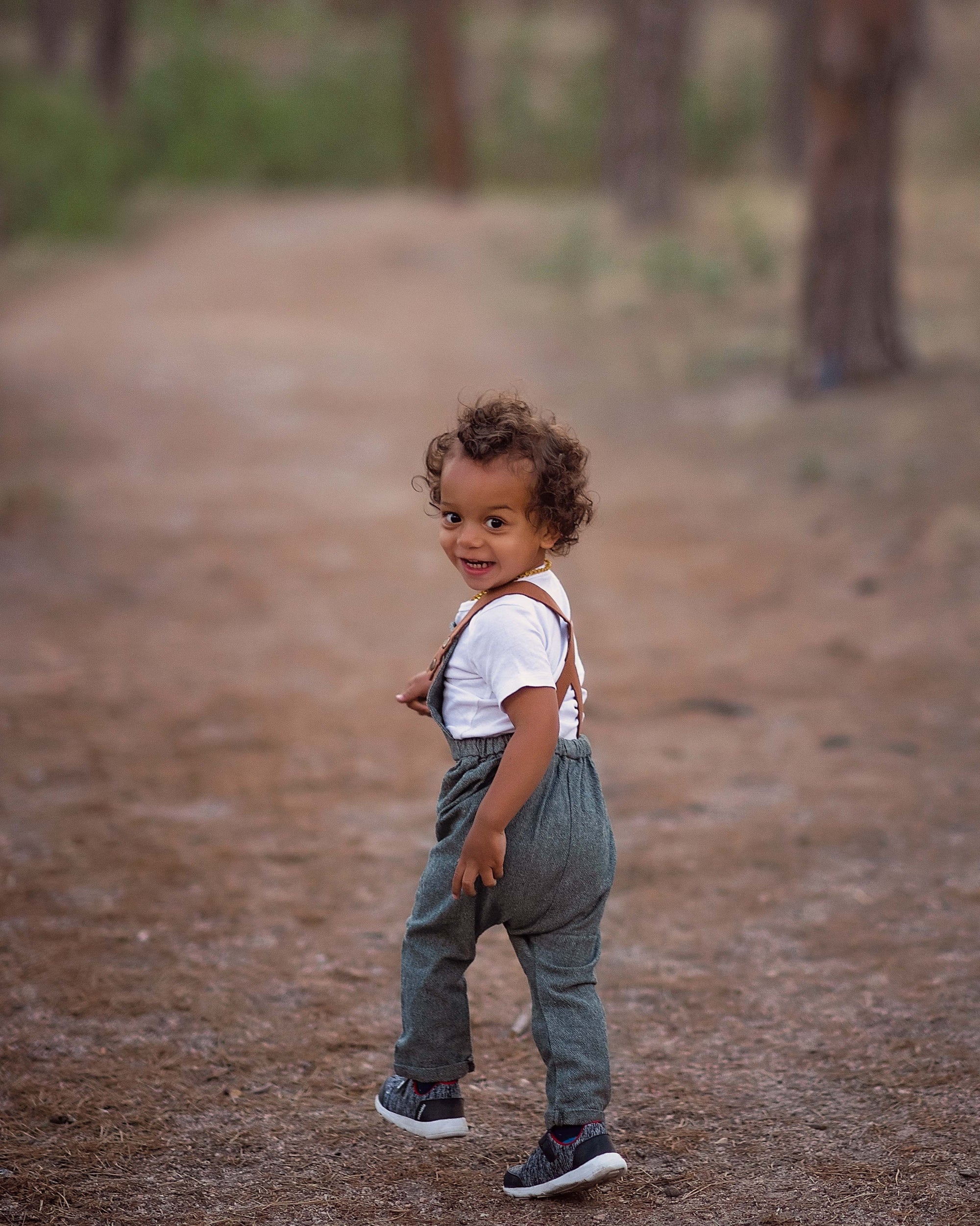 Child wearing suspenders standing on a dirt path with trees in the background