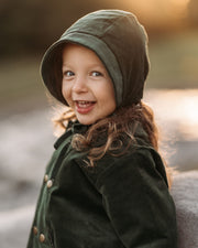 Smiling 3-year-old girl wearing the Evergreen corduroy bonnet and matching coat, highlighting the gender-neutral style and comfortable fit.