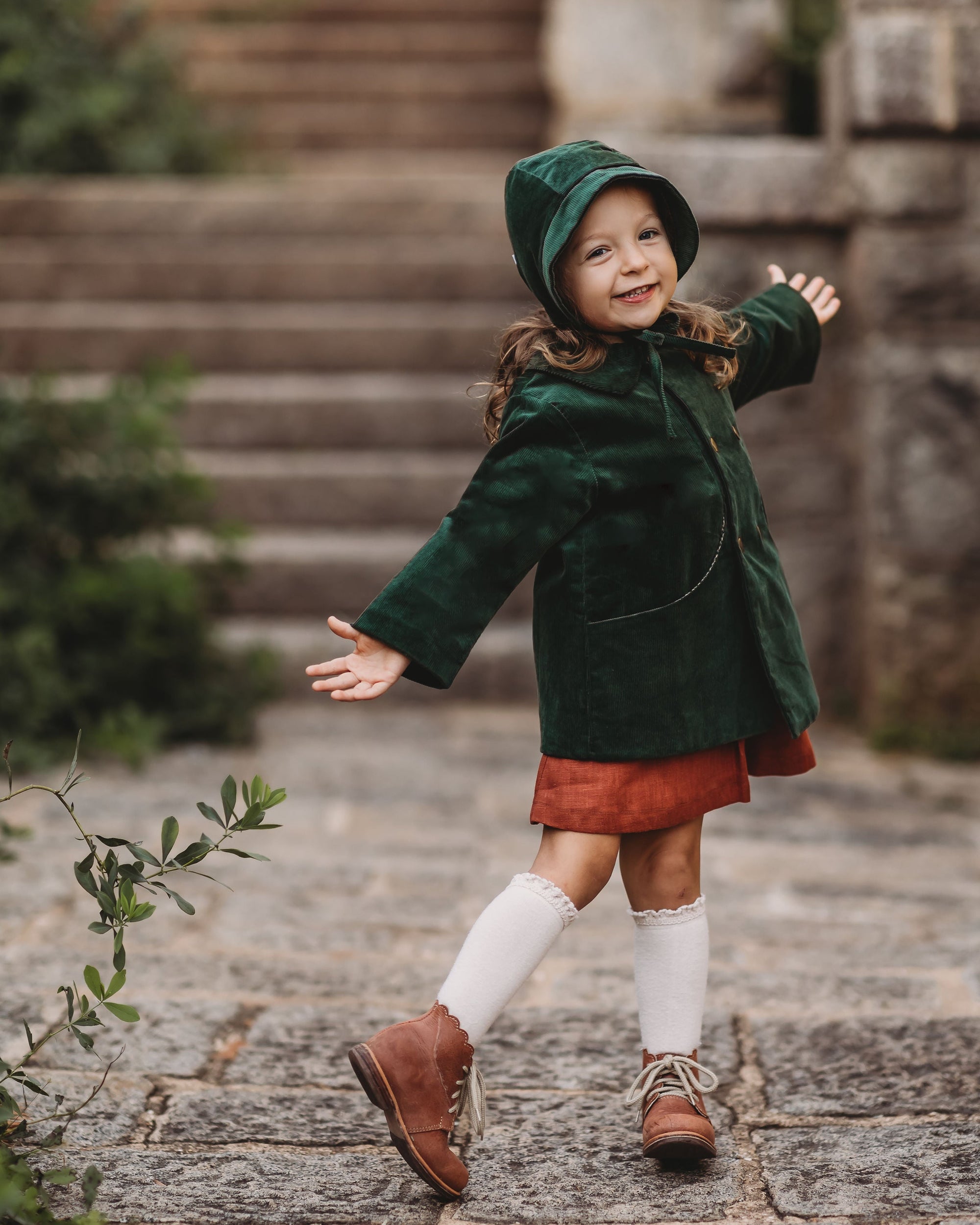 Child wearing a green coat and red skirt standing outdoors on stone steps.
