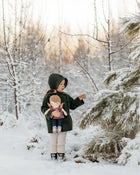 A child standing in a snowy outdoor setting, wearing a dark green hooded coat, beige pants, and black boots, while holding a doll. Snow-covered trees and shrubs fill the background.