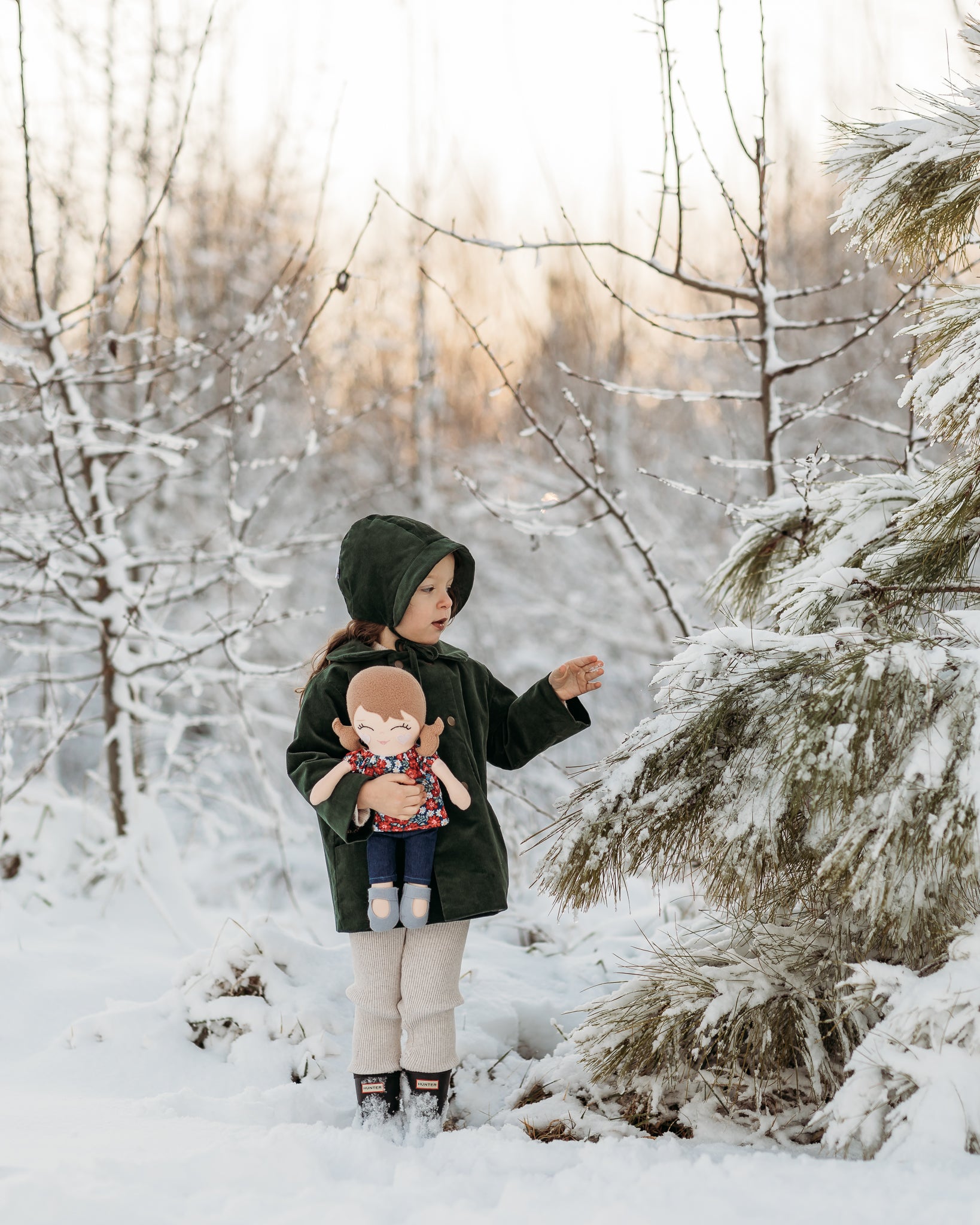 A child standing in a snowy outdoor setting, wearing a dark green hooded coat, beige pants, and black boots, while holding a doll. Snow-covered trees and shrubs fill the background.