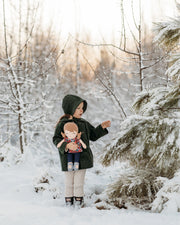 A child standing in a snowy outdoor setting, wearing a dark green hooded coat, beige pants, and black boots, while holding a doll. Snow-covered trees and shrubs fill the background.