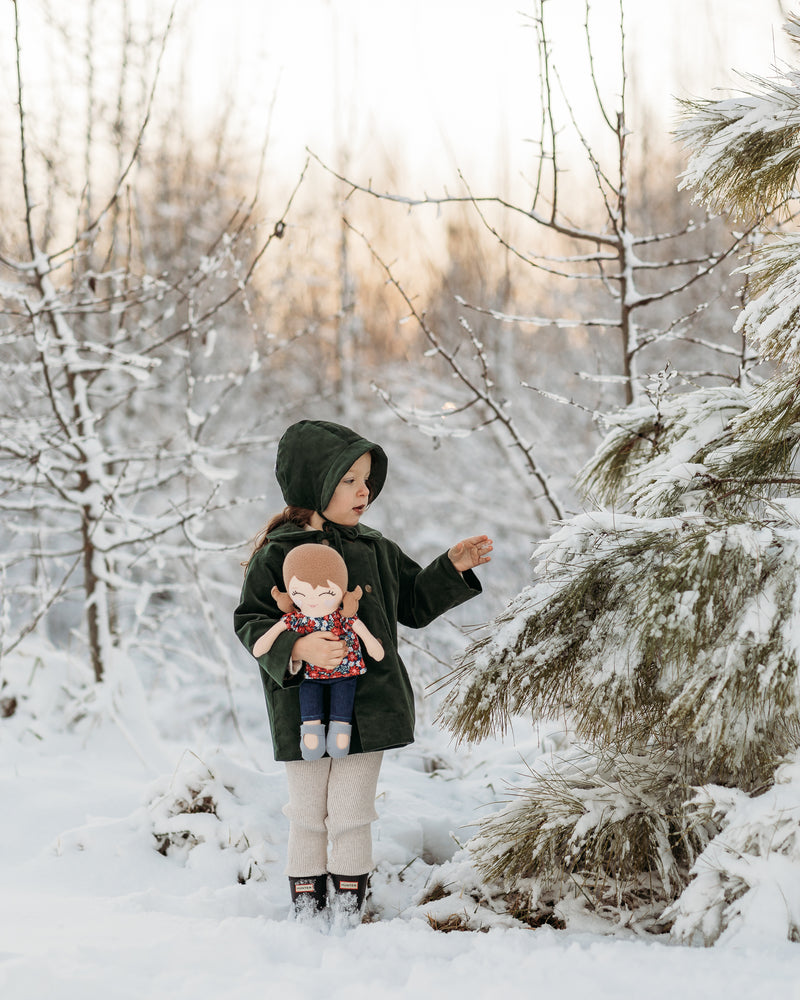 A child standing in a snowy outdoor setting, wearing a dark green hooded coat, beige pants, and black boots, while holding a doll. Snow-covered trees and shrubs fill the background.
