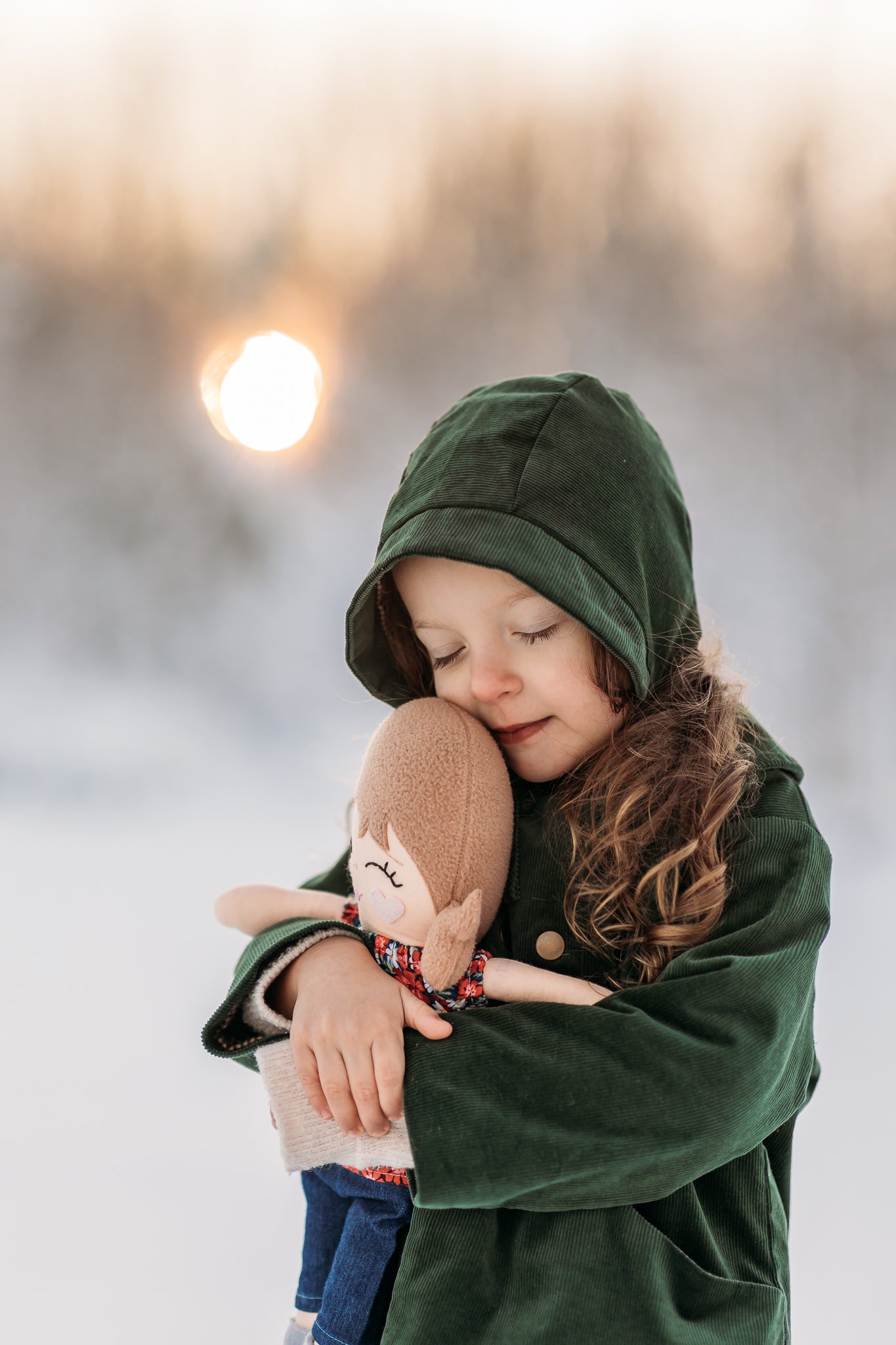 Model girl cuddling a doll in a snowy landscape, wearing the Evergreen bonnet, representing warmth and timeless children’s wear.