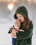 Model girl cuddling a doll in a snowy landscape, wearing the Evergreen bonnet, representing warmth and timeless children’s wear.