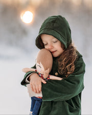 Model girl cuddling a doll in a snowy landscape, wearing the Evergreen bonnet, representing warmth and timeless children’s wear.