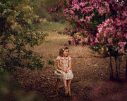 Young girl sitting in a field with flowers wearing a beige blouse with white Swiss dots and a matching beige gingham pinafore. The blouse features a Peter Pan collar with ruffles and short puffy sleeves, while the pinafore showcases a classic gingham pattern, perfect for playful outfits