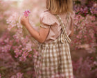 Back details of a young girl wearing a beige blouse with white Swiss dots and a matching beige gingham pinafore. The blouse features a Peter Pan collar with ruffles and short puffy sleeves, while the pinafore showcases a classic gingham pattern and adjustable straps, perfect for playful outfits.