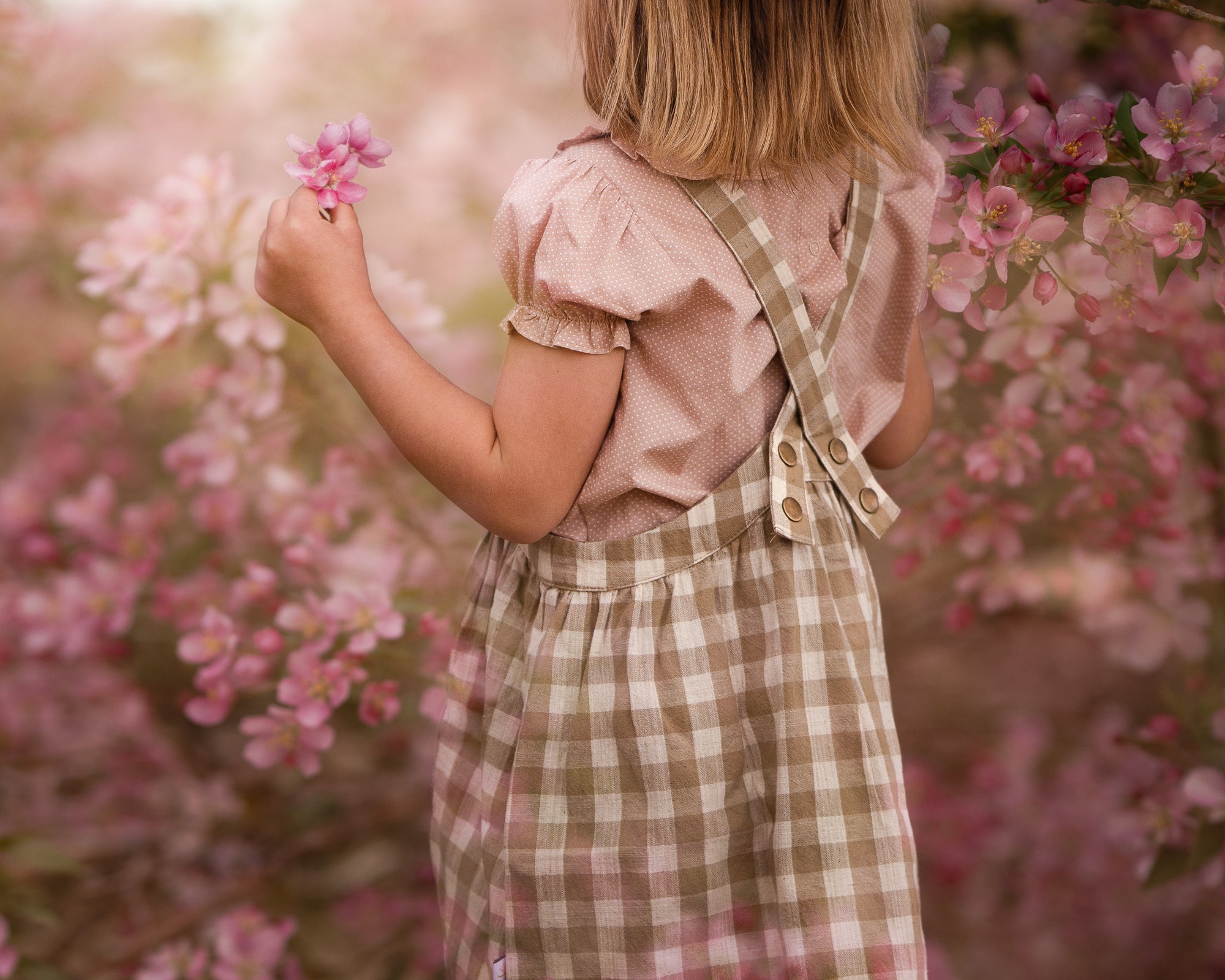 Back details of a young girl wearing a beige blouse with white Swiss dots and a matching beige gingham pinafore. The blouse features a Peter Pan collar with ruffles and short puffy sleeves, while the pinafore showcases a classic gingham pattern and adjustable straps, perfect for playful outfits.