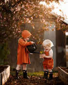 Two children in orange outfits and rain boots playing with a watering can and plant in a garden.