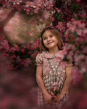 Full view of a young girl wearing a beige blouse with white Swiss dots and a matching beige gingham pinafore. The blouse features a Peter Pan collar with ruffles and short puffy sleeves, while the pinafore showcases a classic gingham pattern, perfect for playful outfits.