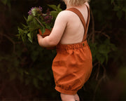 Back view of model boy playing outdoors in Burnt Orange Short Overalls - This image captures another playful moment, emphasizing the overall functionality and stylish appeal of the shortalls for active kids.