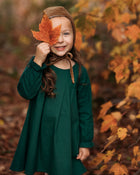 A close-up of a 4-year-old girl holding a leaf to her face while wearing the evergreen linen dress, emphasizing the delicate ruffled hem and fun details.