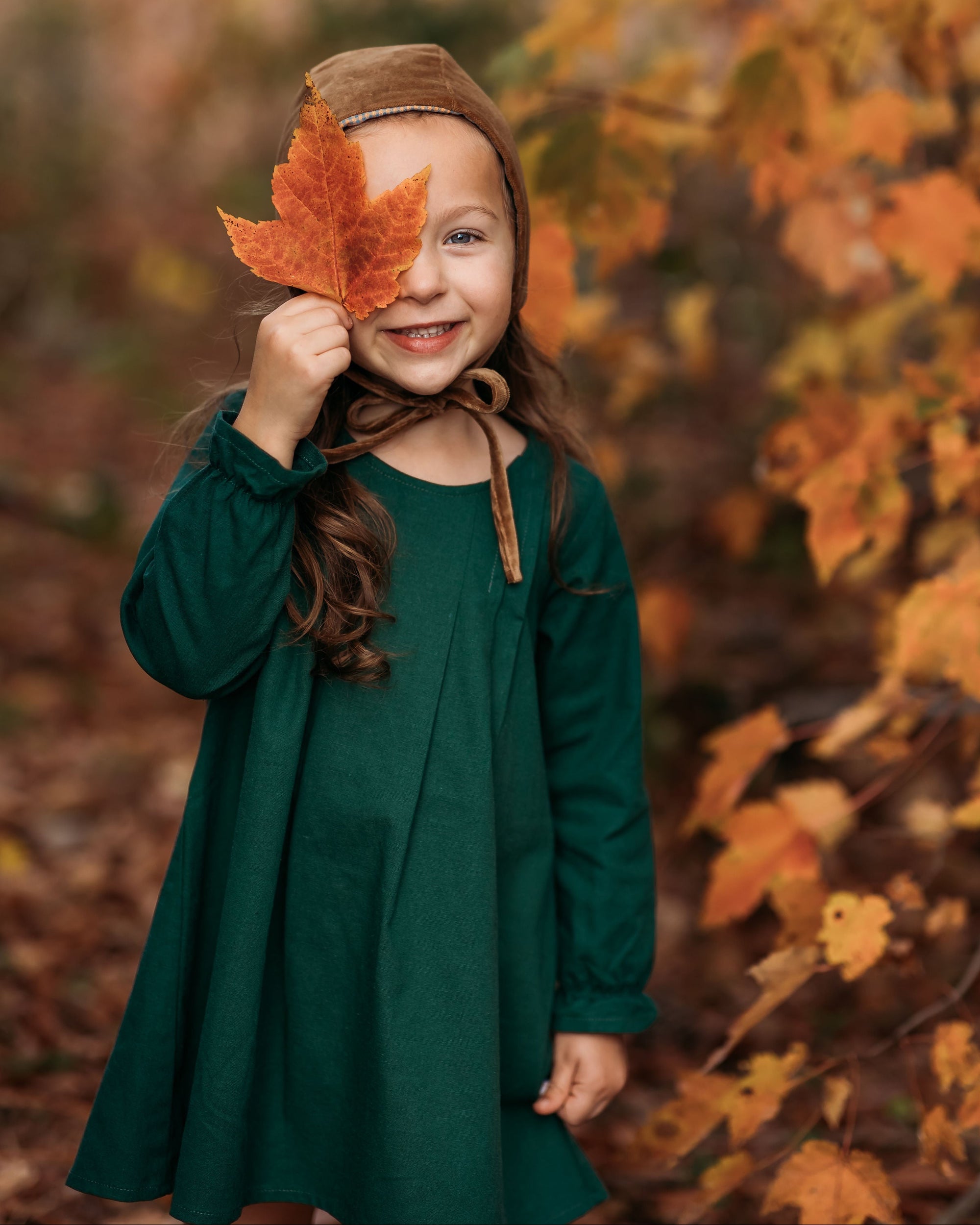 A close-up of a 4-year-old girl holding a leaf to her face while wearing the evergreen linen dress, emphasizing the delicate ruffled hem and fun details.
