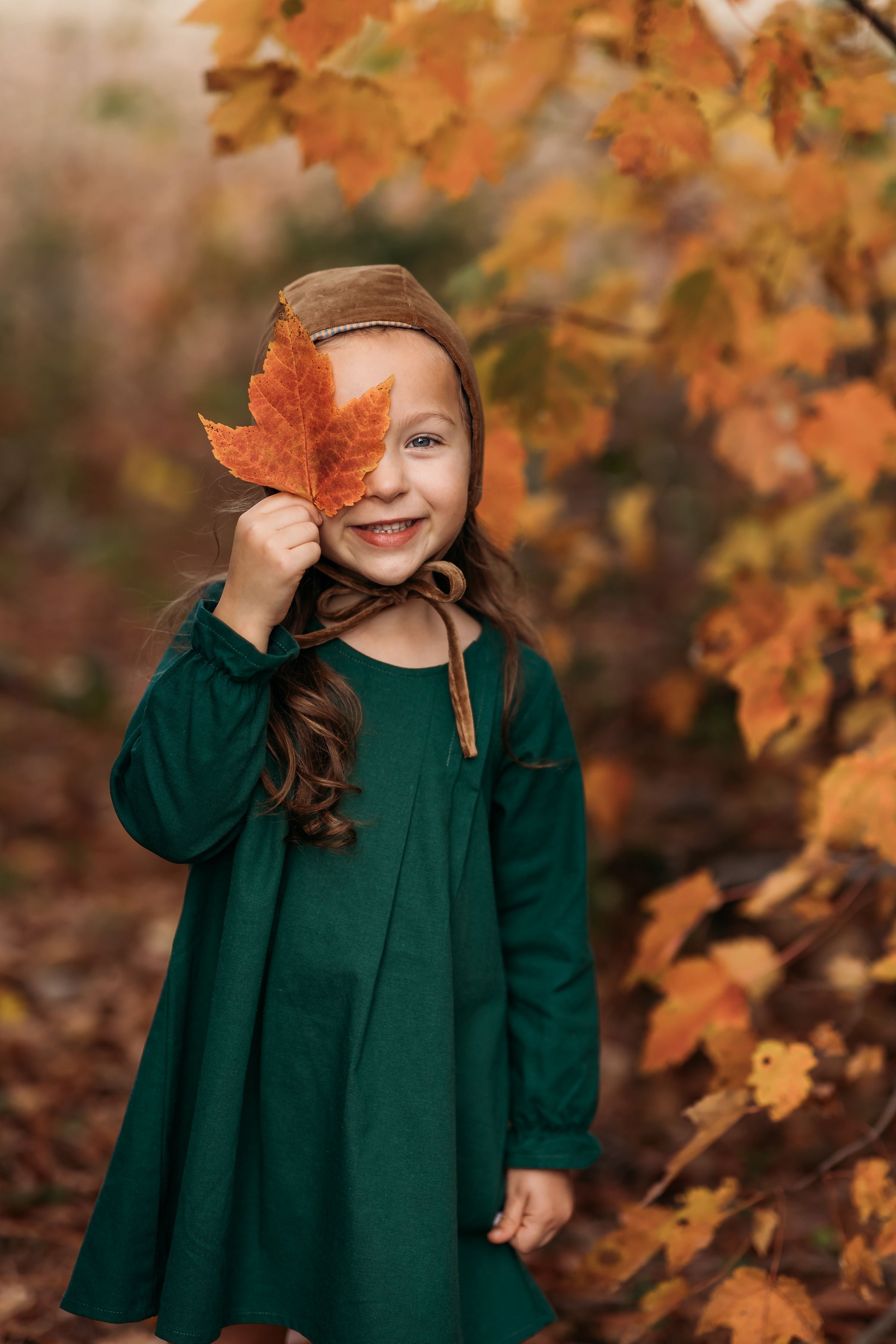 A close-up of a 4-year-old girl holding a leaf to her face while wearing the evergreen linen dress, emphasizing the delicate ruffled hem and fun details.