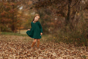 A 4-year-old girl posing sweetly while holding the tip of her dress’s skirt, showcasing the stylish fit and comfortable design of the evergreen linen dress.