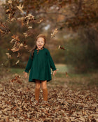 A 4-year-old girl surrounded by colorful autumn leaves, laughing joyfully in her evergreen linen dress, highlighting the playful spirit and flattering A-line shape.