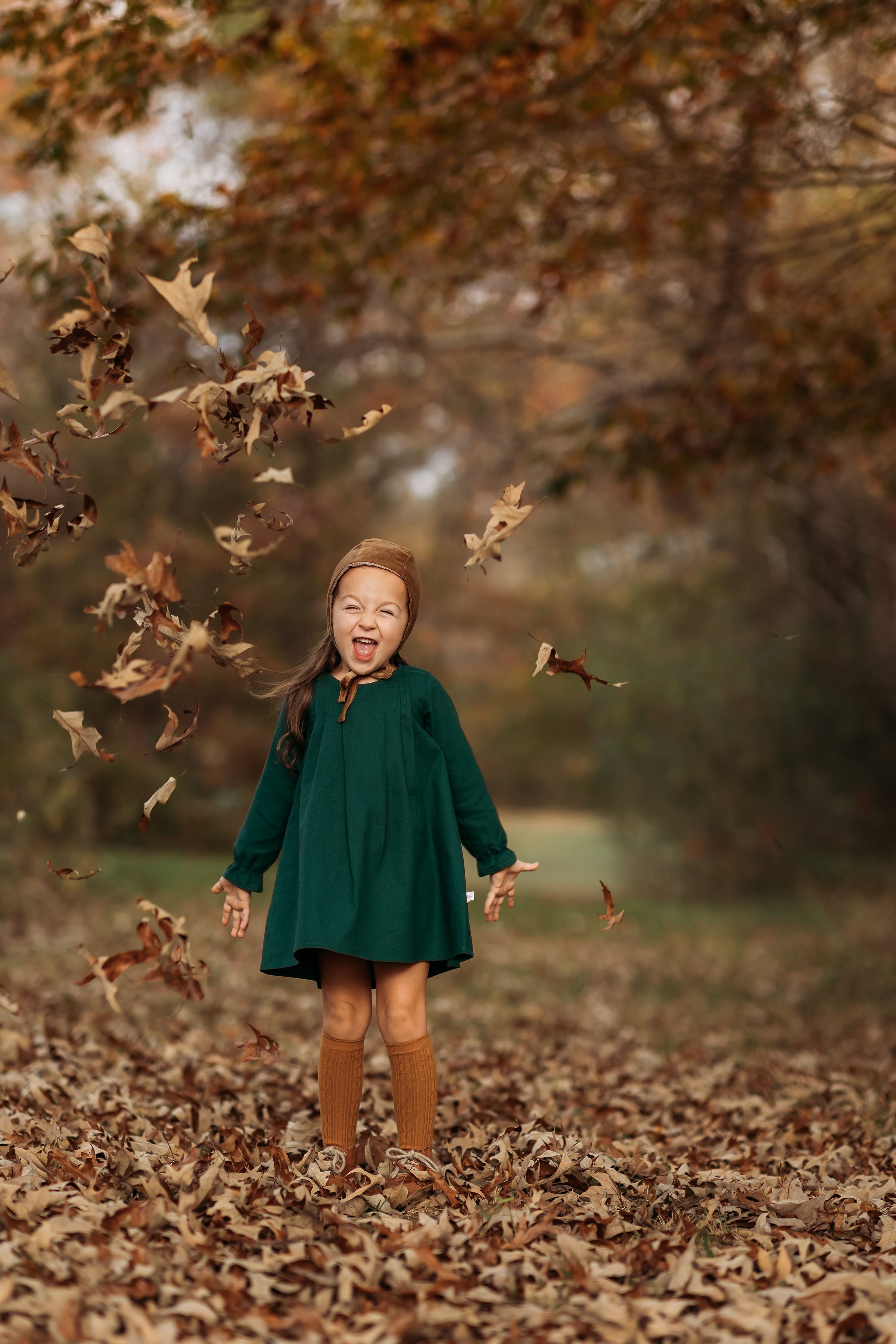 A 4-year-old girl surrounded by colorful autumn leaves, laughing joyfully in her evergreen linen dress, highlighting the playful spirit and flattering A-line shape.
