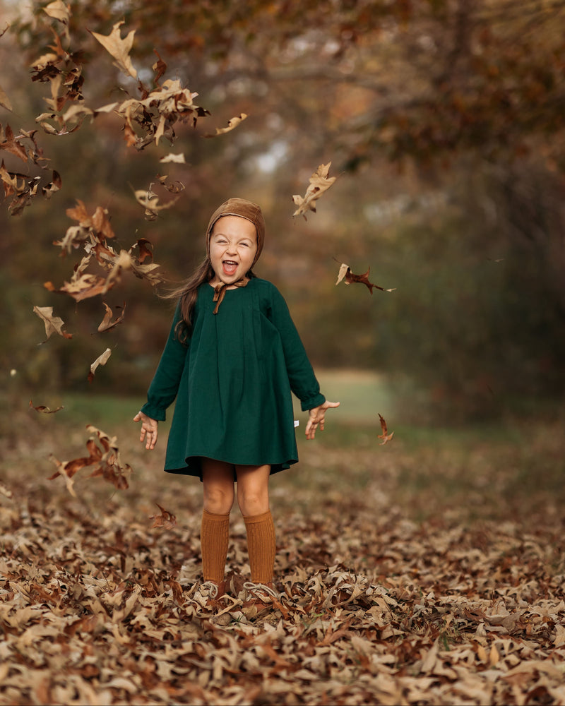 A 4-year-old girl surrounded by colorful autumn leaves, laughing joyfully in her evergreen linen dress, highlighting the playful spirit and flattering A-line shape.