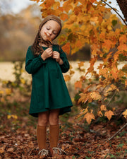 A 4-year-old girl standing in a fall setting, wearing the evergreen linen dress while holding a brown leaf, capturing the seasonal charm and style of the outfit.